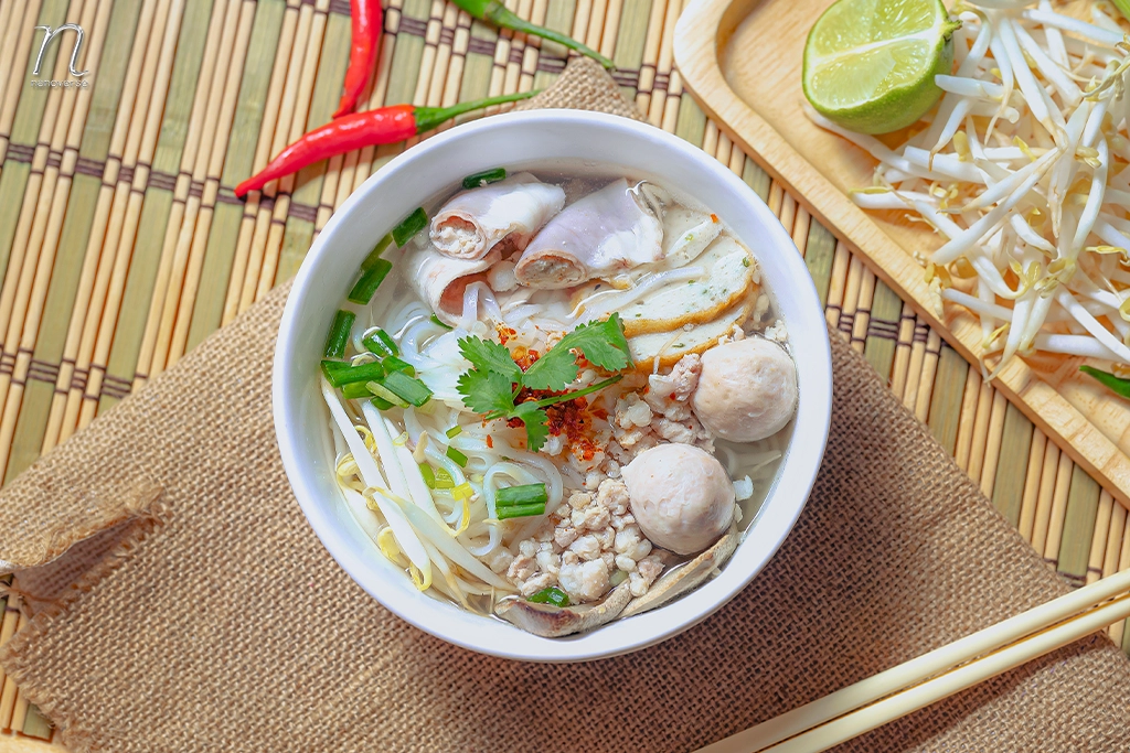 ชามก๋วยเตี๋ยวไก่เสิร์ฟพร้อมลูกชิ้น ถั่วงอก ต้นหอม ผักชี และพริก อยู่บนเสื่อไม้ไผ่ มีมะนาวและถั่วงอกวางอยู่ข้างๆ